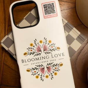 May include: White phone case with a black border, featuring a floral design with pink and yellow flowers, and the text "BLOOMING LOVE ESTABLISHED 2025." A QR code sticker is in the upper right corner with the text "SCAN HERE!"