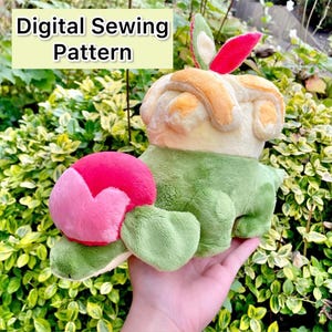 May include: A plush toy resembling a green and yellow creature with a pink heart-shaped head. The toy is made of soft, fuzzy fabric and has a friendly expression. The text "Digital Sewing Pattern" is displayed above the toy.