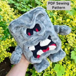 May include: A plush, gray, square-shaped character with a grumpy expression, featuring red eyes, a wide mouth with white teeth, and small arms and legs. The text "PDF Sewing Pattern" is in a white box.