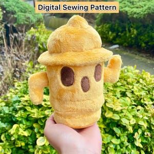 May include: A plush toy shaped like a fire hydrant. The toy is yellow with brown eyes and a brown mouth. The toy is being held in a person's hand. The text "Digital Sewing Pattern" is displayed above the toy.