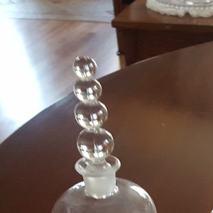 May include: A clear glass bottle with a round body and a decorative stopper. The stopper is made of clear glass and has a series of round beads stacked on top of each other. The bottle has a round base with a series of small, round beads.