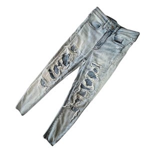 May include: Light blue denim jeans with distressed detailing. The jeans feature multiple large rips and tears across the front of the legs, with visible patches underneath. The jeans have a classic five-pocket design and a button and zipper closure.