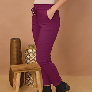 Pode incluir: Calças roxo-profundo com cintura alta elástica, cordão funcional, bolsos laterais e bainhas dobradas. As calças são combinadas com uma blusa de manga curta em tom neutro e botas de tornozelo pretas com saltos em bloco.