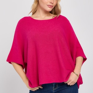 May include: A vibrant fuchsia pink knit poncho top featuring a wide boat neckline and loose, short dolman sleeves. The garment has a relaxed, boxy silhouette that drapes comfortably over the torso, paired with dark blue denim jeans.