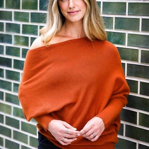 May include: A burnt orange knit jumper featuring an asymmetrical off-the-shoulder neckline, long sleeves with fitted cuffs, and a relaxed, draped silhouette that falls loosely over the torso, paired with simple black trousers.