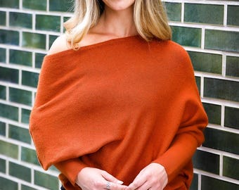 May include: A burnt orange knit jumper featuring an asymmetrical off-the-shoulder neckline, long sleeves with fitted cuffs, and a relaxed, draped silhouette that falls loosely over the torso, paired with simple black trousers.