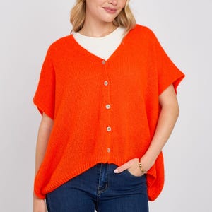 Puede incluir: Cárdigan naranja brillante de manga corta con cuello en V y cierre de botones. El cárdigan se lleva sobre una camiseta blanca y se combina con vaqueros azules. Se ve una pulsera dorada en la muñeca.