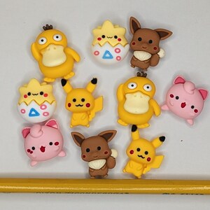 Puede incluir: Una colecci&oacute;n de adornos coloridos con forma de personajes de dibujos animados, incluyendo patos amarillos, Pikachu y criaturas rosas. Un l&aacute;piz amarillo est&aacute; en la parte inferior de la imagen. Probablemente sean para manualidades o decoraci&oacute;n.