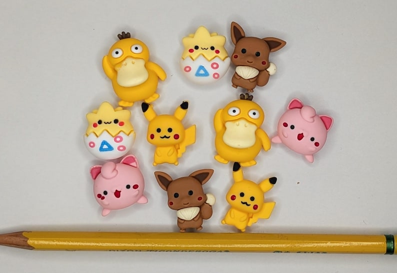 Puede incluir: Una colecci&oacute;n de adornos coloridos con forma de personajes de dibujos animados, incluyendo patos amarillos, Pikachu y criaturas rosas. Un l&aacute;piz amarillo est&aacute; en la parte inferior de la imagen. Probablemente sean para manualidades o decoraci&oacute;n.