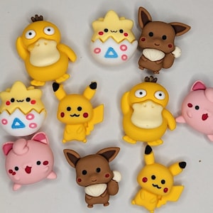 Puede incluir: Una colecci&oacute;n de adornos coloridos con forma de personajes. El surtido incluye figuras amarillas con forma de pato, figuras Pikachu amarillas, figuras Eevee marrones, figuras rosas y figuras con forma de huevo blanco y amarillo. Estas piezas decorativas tienen detalles en negro, rojo y azul.