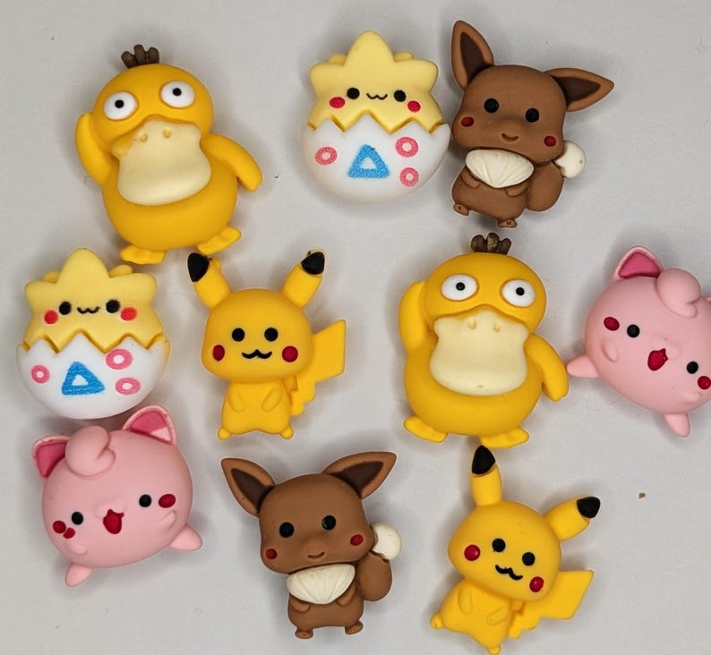 Puede incluir: Una colecci&oacute;n de adornos coloridos con forma de personajes. El surtido incluye figuras amarillas con forma de pato, figuras Pikachu amarillas, figuras Eevee marrones, figuras rosas y figuras con forma de huevo blanco y amarillo. Estas piezas decorativas tienen detalles en negro, rojo y azul.