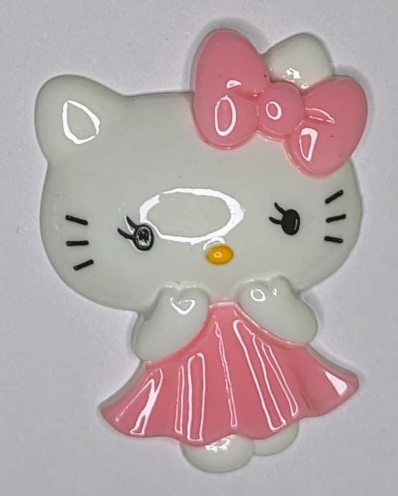 May include: A white ceramic figurine of a cartoon cat wearing a pink dress and a pink bow. The cat has black eyes and a yellow nose.