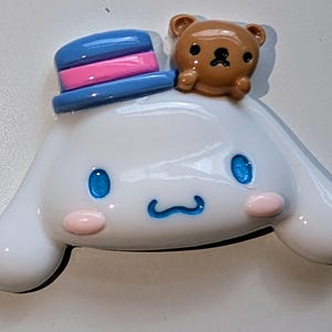 May include: A white ceramic figurine of a dog wearing a blue and pink hat with a brown bear on top. The dog has blue eyes and a pink nose.