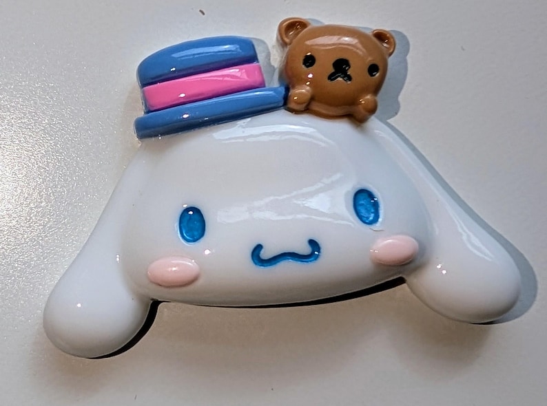 May include: A white ceramic figurine of a dog wearing a blue and pink hat with a brown bear on top. The dog has blue eyes and a pink nose.