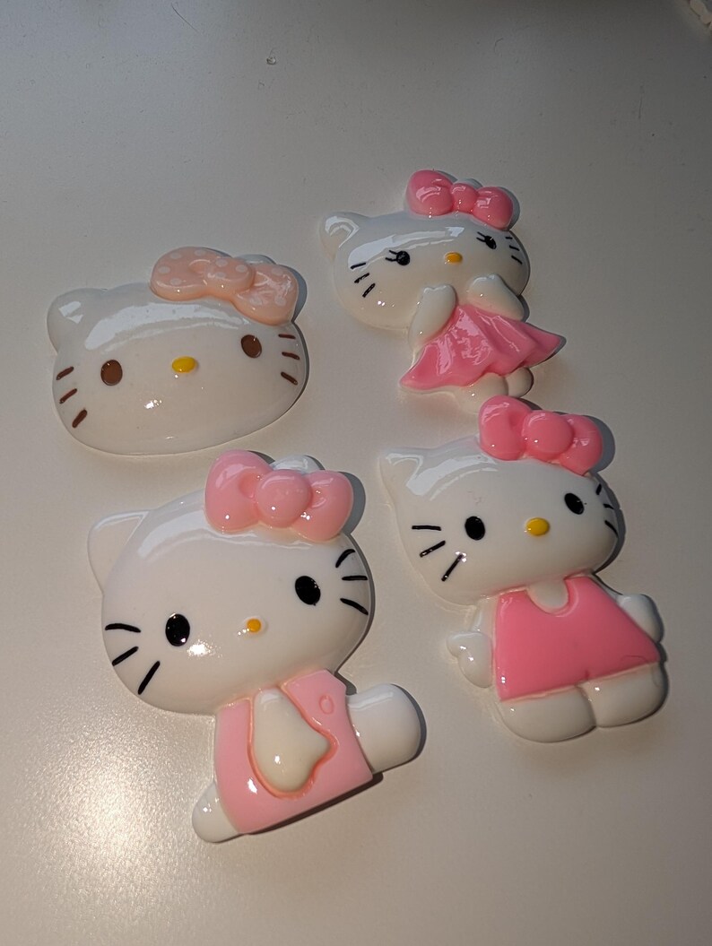 May include: Four white Hello Kitty figurines with pink bows and dresses. The figurines are made of plastic and have a glossy finish.