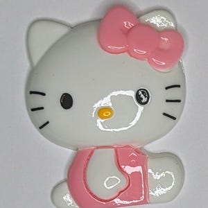 May include: A white and pink Hello Kitty character figurine with a pink bow on its head. The figurine is wearing a pink dress and has black eyes and a yellow nose.