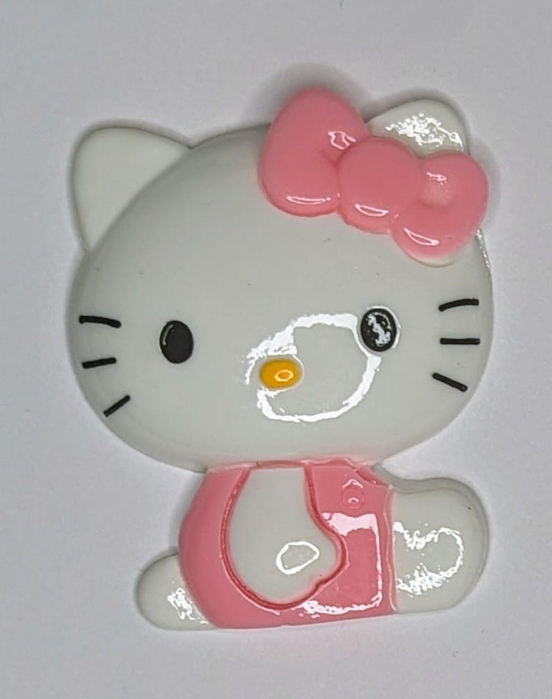 May include: A white and pink Hello Kitty character figurine with a pink bow on its head. The figurine is wearing a pink dress and has black eyes and a yellow nose.