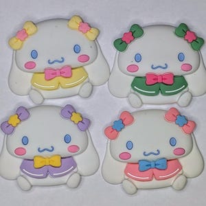 Puede incluir: Cuatro peluches blancos con forma de perro con lazos y atuendos de diferentes colores. Los perros tienen mejillas rosadas, ojos azules y un bigote negro. Los atuendos son amarillos, verdes, morados y rosas.