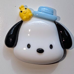 May include: A white ceramic figurine of a dog wearing a blue hat with a yellow chick on top. The dog has black ears and a black nose.