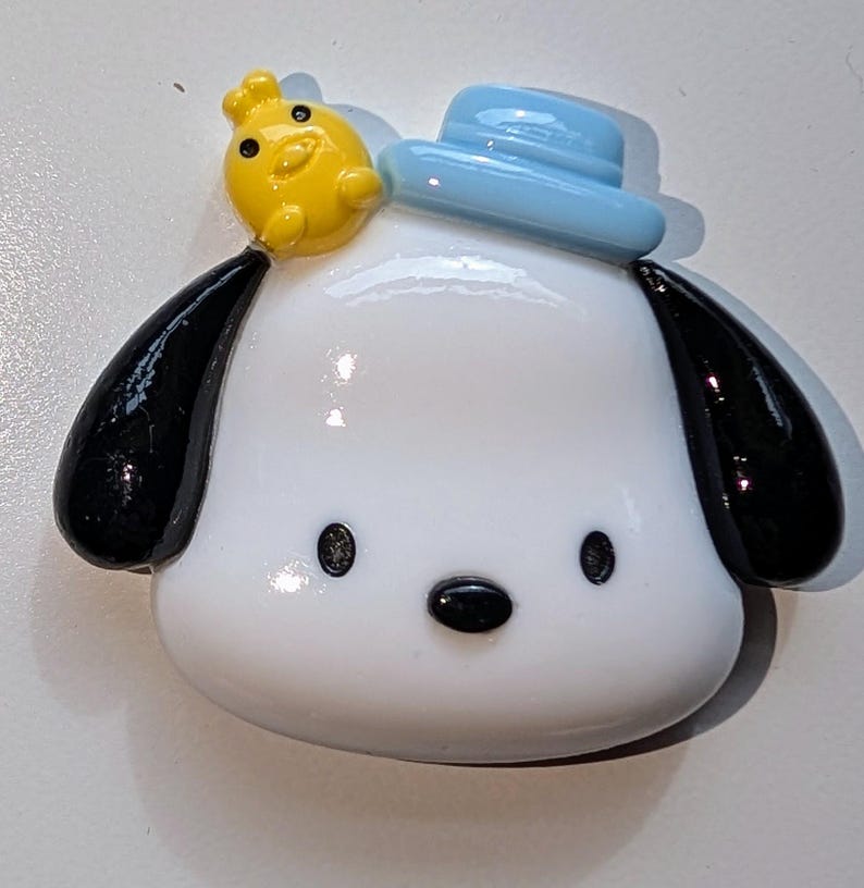 May include: A white ceramic figurine of a dog wearing a blue hat with a yellow chick on top. The dog has black ears and a black nose.