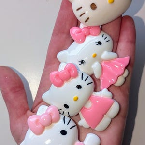 May include: Four white Hello Kitty figurines with pink bows and dresses. The figurines are different sizes and are arranged in a stack.