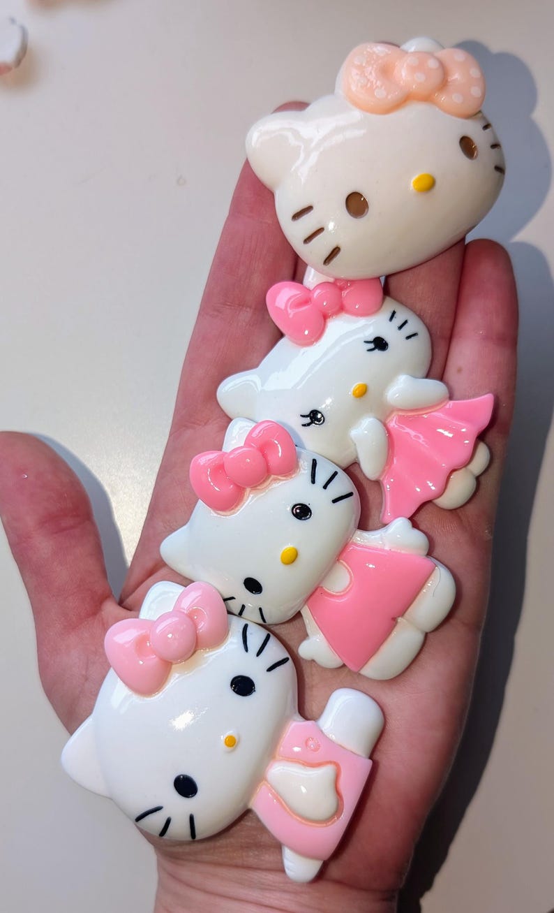 May include: Four white Hello Kitty figurines with pink bows and dresses. The figurines are different sizes and are arranged in a stack.