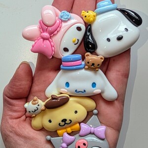 May include: Five colorful cartoon character charms with different expressions and accessories. The charms are made of resin and have a glossy finish. The characters include a pink bunny with a bow, a white dog with a blue hat, a white dog with a blue hat and a brown bear, a yellow dog with a brown hat, and a gray cat with a purple bow.