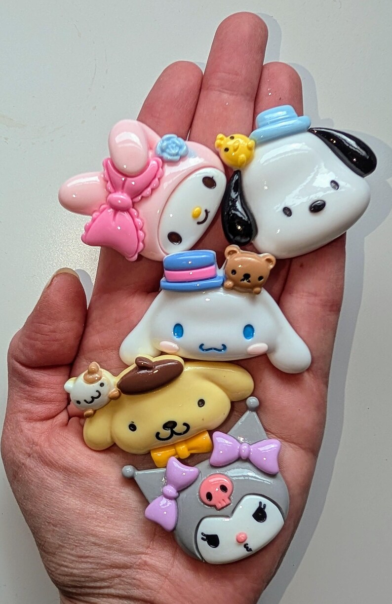 May include: Five colorful cartoon character charms with different expressions and accessories. The charms are made of resin and have a glossy finish. The characters include a pink bunny with a bow, a white dog with a blue hat, a white dog with a blue hat and a brown bear, a yellow dog with a brown hat, and a gray cat with a purple bow.