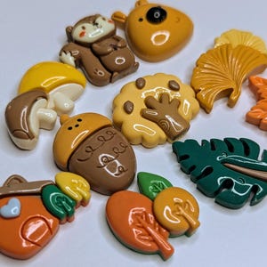 May include: A set of 10 colorful clay charms featuring autumnal themes. The charms include a squirrel, mushroom, acorn, tree, leaves, and a bear. These charms are perfect for adding a touch of fall to your crafts.