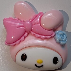 May include: A pink and white Hello Kitty character charm with a pink bow and a blue flower. The charm is made of plastic and has a glossy finish.