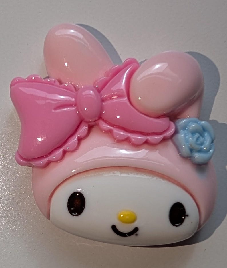 May include: A pink and white Hello Kitty character charm with a pink bow and a blue flower. The charm is made of plastic and has a glossy finish.