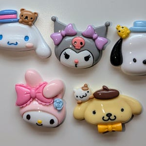 May include: Six colorful character charms featuring popular cartoon characters. The charms are made of plastic and have a glossy finish. The characters include a pink and white bunny with a pink bow, a gray and white cat with purple bows, a white dog with a blue hat, a white dog with a brown hat, a yellow dog with a brown hat, and a brown bear wearing a blue hat.