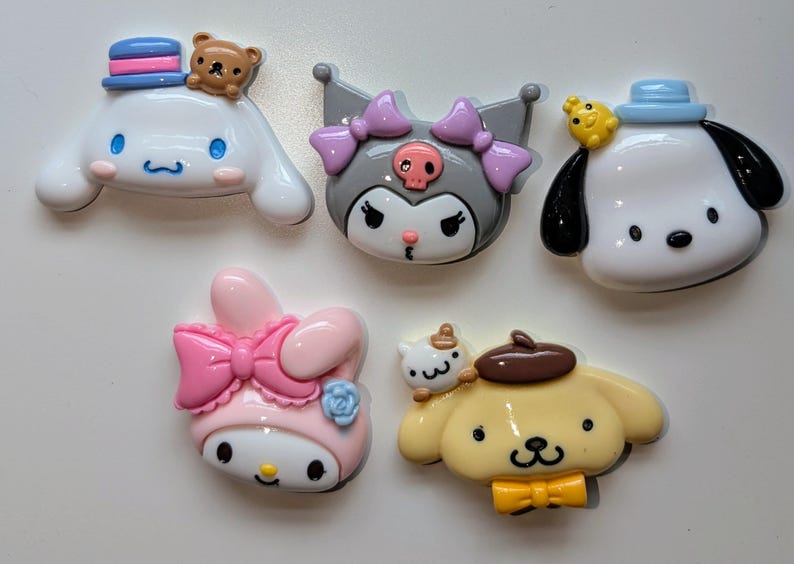 May include: Six colorful character charms featuring popular cartoon characters. The charms are made of plastic and have a glossy finish. The characters include a pink and white bunny with a pink bow, a gray and white cat with purple bows, a white dog with a blue hat, a white dog with a brown hat, a yellow dog with a brown hat, and a brown bear wearing a blue hat.