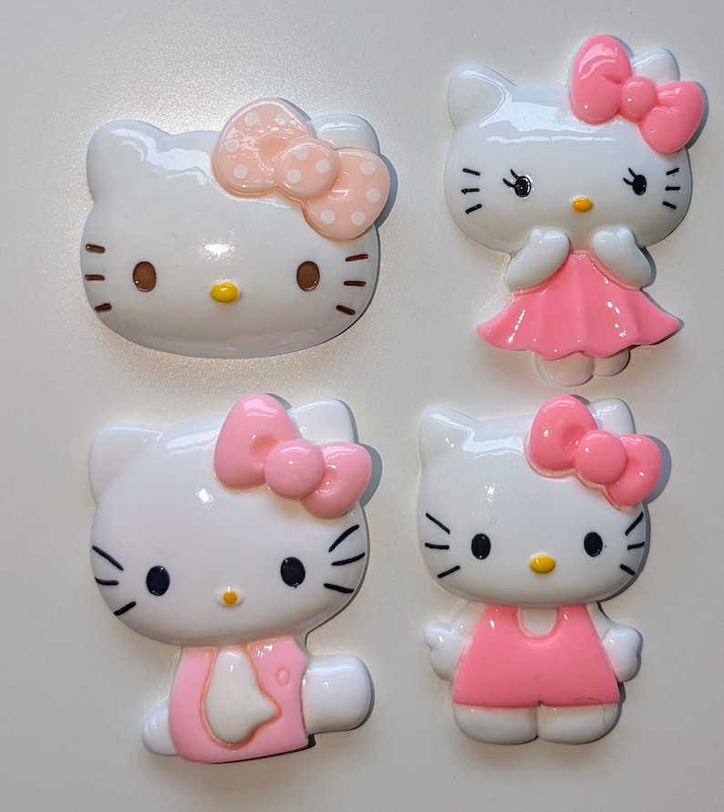 May include: Four white Hello Kitty resin charms with pink bows and dresses. The charms are in different poses, with one charm featuring a pink bow with white polka dots.