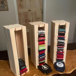 May include: Three wooden display stands with a natural finish. Each stand is filled with folded beverage insulators in various colors. Some insulators are black and navy blue with white logos. The stands are on a gold-colored table runner.