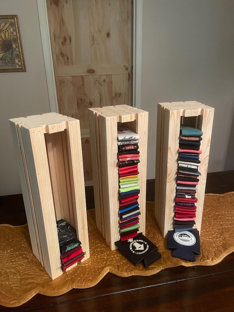 May include: Three wooden display stands with a natural finish. Each stand is filled with folded beverage insulators in various colors. Some insulators are black and navy blue with white logos. The stands are on a gold-colored table runner.