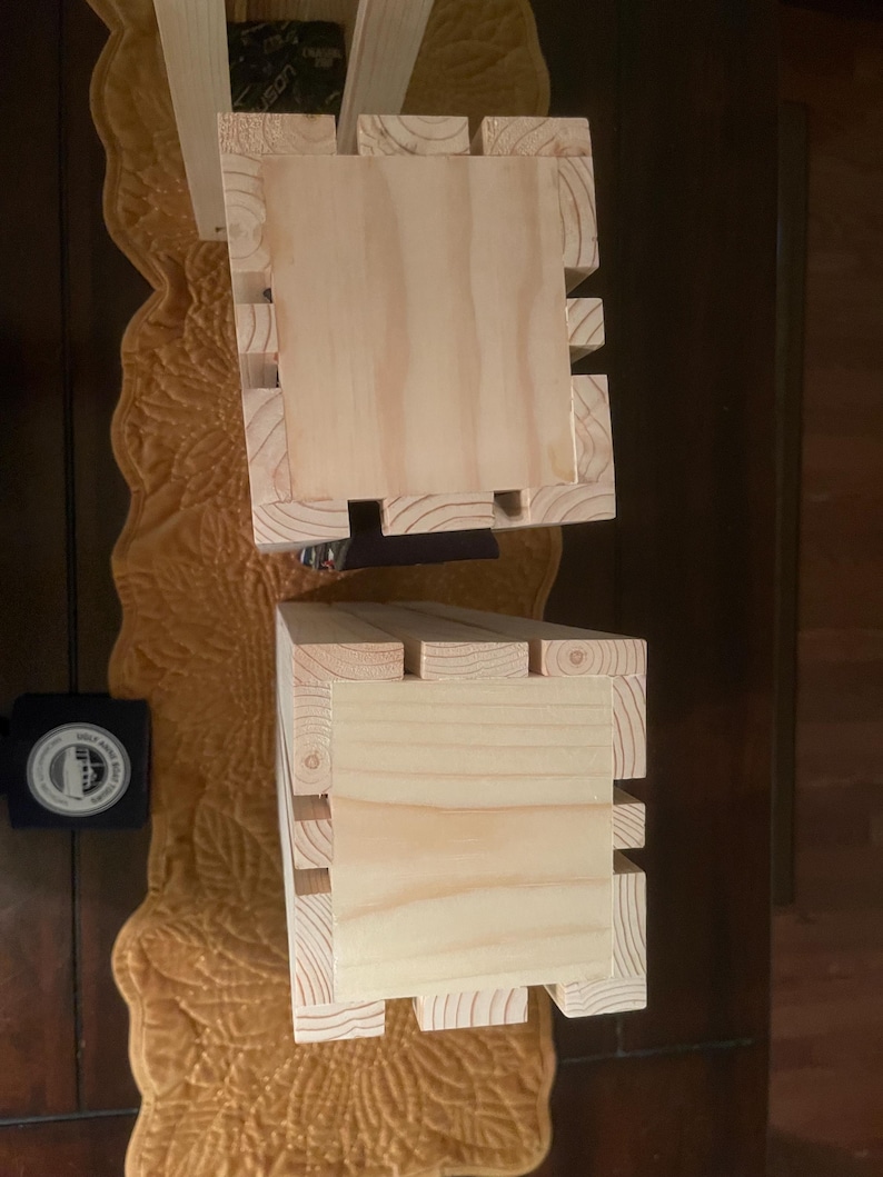 May include: Two unfinished wooden boxes with a natural wood grain. The boxes are rectangular with a square top and a slotted design on the sides. The boxes are likely for storage or display.