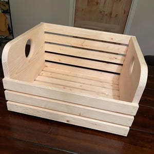 May include: A natural wood storage crate with two cut-out handles on the sides. The crate is made of unfinished pine wood and has a rectangular shape.