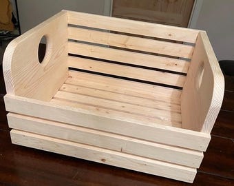 May include: A natural wood storage crate with two cut-out handles on the sides. The crate is made of unfinished pine wood and has a rectangular shape.
