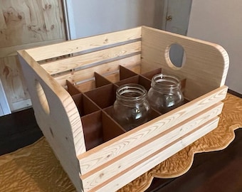 May include: A wooden crate with a light finish, featuring a divided interior and two round handle cutouts. Two clear glass jars are visible inside. The crate is sitting on a gold-colored runner.