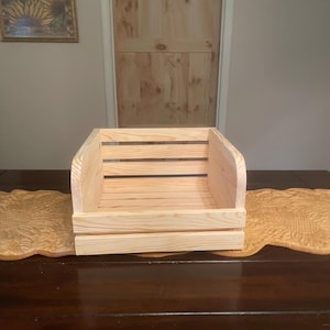 May include: A light-colored wooden crate with a slatted design, set on a dark wooden table. The crate has a rectangular shape with curved sides. A framed sunflower painting hangs on the wall in the background.