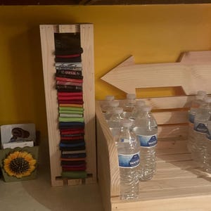May include: A wooden rack displays folded shirts in various colors, alongside a wooden crate filled with bottled water. A wooden arrow points right, and a sunflower-themed container sits nearby.
