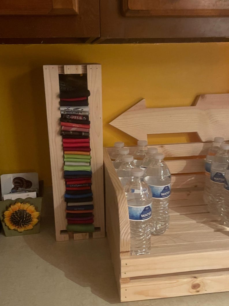 May include: A wooden rack displays folded shirts in various colors, alongside a wooden crate filled with bottled water. A wooden arrow points right, and a sunflower-themed container sits nearby.