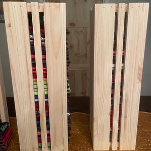 May include: Two unfinished wooden storage units with vertical slats, filled with colorful fabric rolls. The units are made of light-colored wood and sit on a gold-colored surface. The fabric rolls are various colors and patterns.