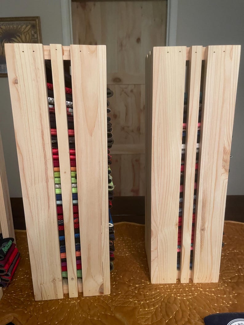 May include: Two unfinished wooden storage units with vertical slats, filled with colorful fabric rolls. The units are made of light-colored wood and sit on a gold-colored surface. The fabric rolls are various colors and patterns.