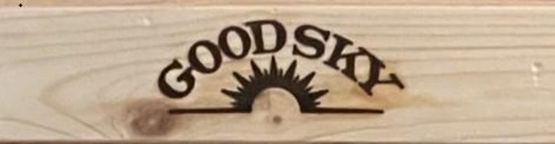 May include: A wooden sign with the words "GOOD SKY" in a dark brown, arched font. Below the text is a stylized sun design. The sign is made of light-colored wood with visible grain.