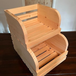 Puede incluir: Una caja de almacenamiento de madera de dos niveles. La caja está construida con madera de color claro y presenta una base de listones y paneles laterales curvos. El diseño es simple y funcional, adecuado para diversas necesidades de almacenamiento. La caja es probablemente hecha a mano.
