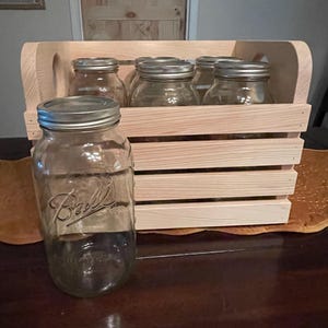 May include: A wooden crate with a handle holds five glass Ball jars with silver lids. A larger Ball jar sits in front of the crate. The jars are clear, and the crate is made of light-colored wood.