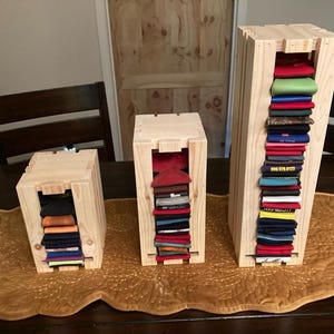 May include: Three wooden storage crates of varying sizes, each with a rectangular opening revealing folded clothing. The crates are made of light-colored wood and sit on a brown table runner. The folded items are in various colors.