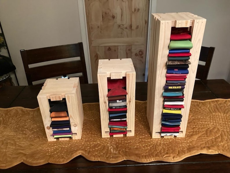 May include: Three wooden storage crates of varying sizes, each with a rectangular opening revealing folded clothing. The crates are made of light-colored wood and sit on a brown table runner. The folded items are in various colors.
